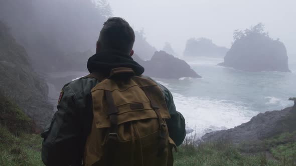 Guy in Pilot Jacket Traveling By Green Mountain to Rocky Coastline Back View