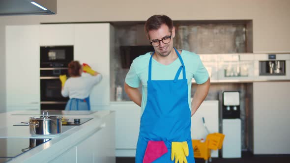 Man Janitor Putting on Apron Preparing for Work in Kitchen with Colleague Cleaning on Background