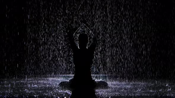 A Silhouette Looms Under the Streams of Rain with Folded Hands in a Lotus Position. Black Background