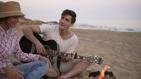 Young Man is Playing Guitar By the Fire Sitting on the Beach Together with Friends