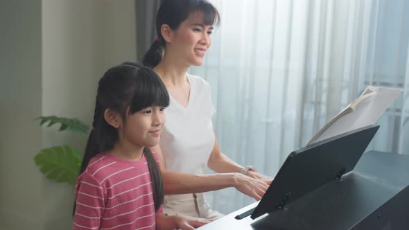 Asian happy mother teach young child daughter enjoy playing piano together in living room at home.