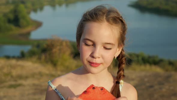 Little Girl with Plait Bites Juicy Watermelon on Riverbank