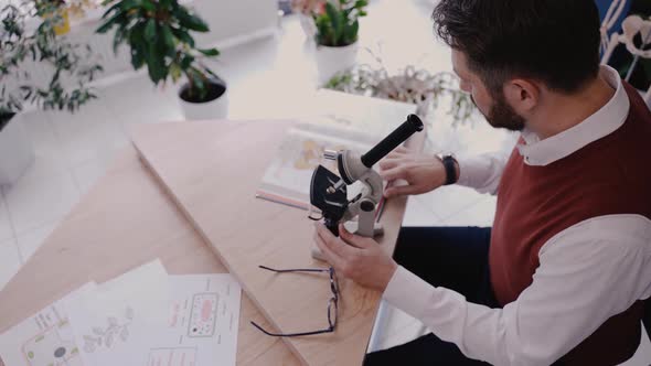 Male Teacher Using Microscope and Looking at Biology Textbook in Modern School