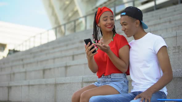 Couple of Mixed-Race Teenagers Sitting Outdoor Scrolling Web Pages on Smartphone
