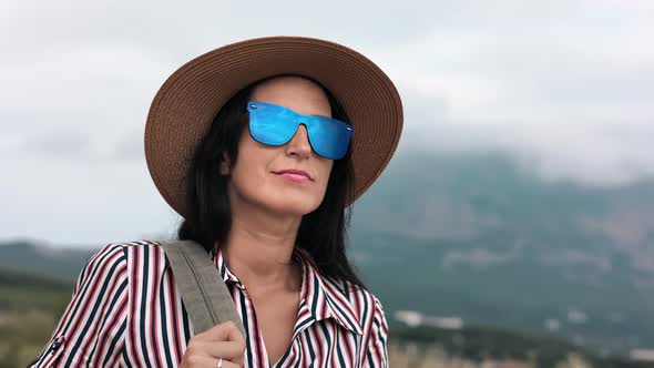 Face Travel Woman in Sunglasses and Straw Hat Putting on Backpack at Dramatic Mountain Sea Landscape