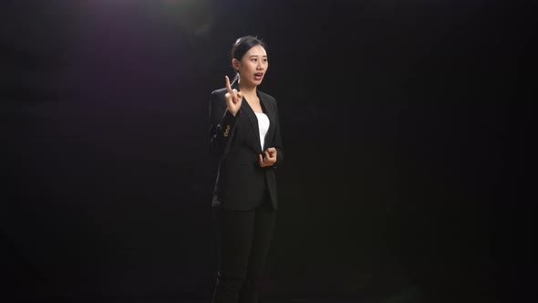 Asian Speaker Woman In Business Suit Holding Her Hands Together While Speaking In The Black Studio