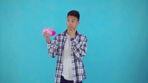 Thoughtful Young Asian Man Holding a Piggy Bank on Blue Background