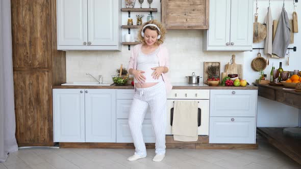 Happy Pregnant Woman Wearing Headphones Dancing In Kitchen.