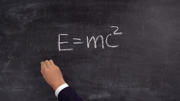 The teacher's hand writes the formula E=mc2 on the blackboard