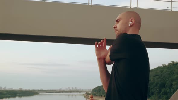 Man Runner in Black Sport Uniform Stretching Hands Before Sports on Pedestrian Bridge at Dawn