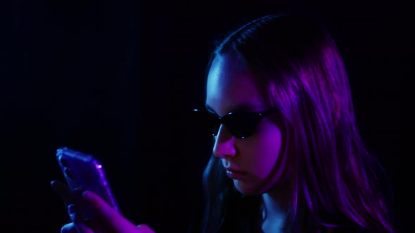 Side View Serious Teenage Girl in Black Sunglasses Browsing Mobile Phone Under Violet Neon Light on