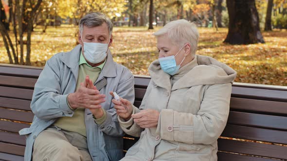 Elderly Couple in Medical Masks in Autumn Park Spouses Disinfect Hands with Antiseptic Sitting on