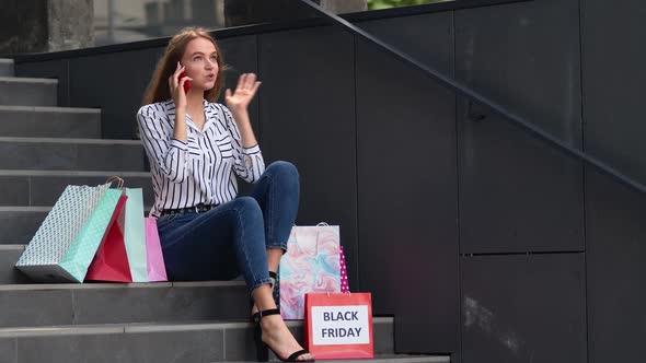 Girl Sitting on Stairs with Bags Talking on Mobile Phone About Sale in Shopping Mall in Black Friday