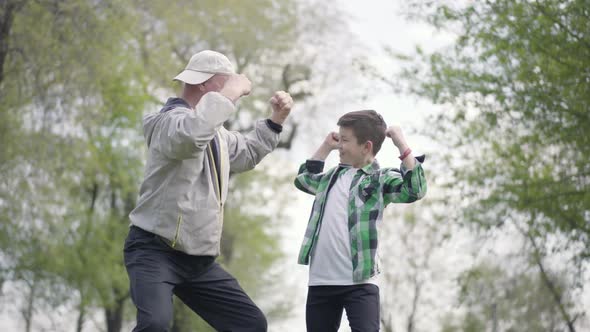 The Little Boy in a Checkered Shirt and Positive Old Man Showing Each Other Muscles in the Park