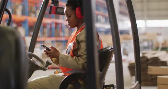 African american female worker wearing safety suit and using smartphone in warehouse