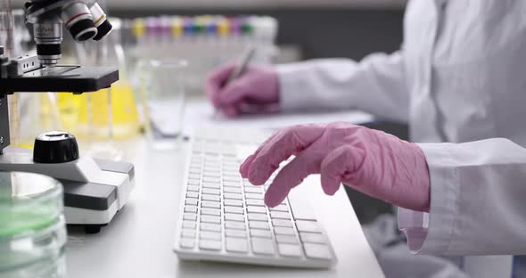 Scientist in Gloves Works on Keyboard at the Desktop and Writes Notes