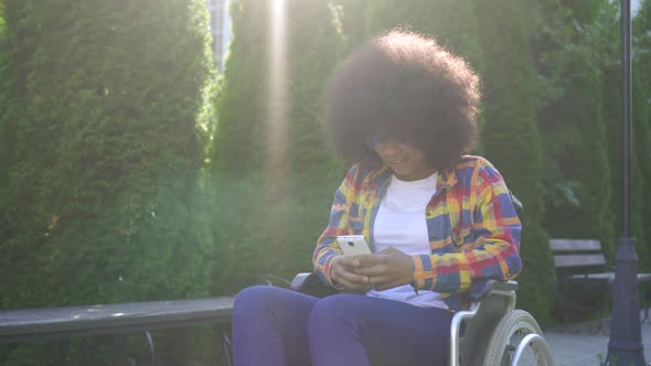 African American Woman with an Afro Hairstyle Disabled in a Wheelchair Uses a Smartphone Sunflare