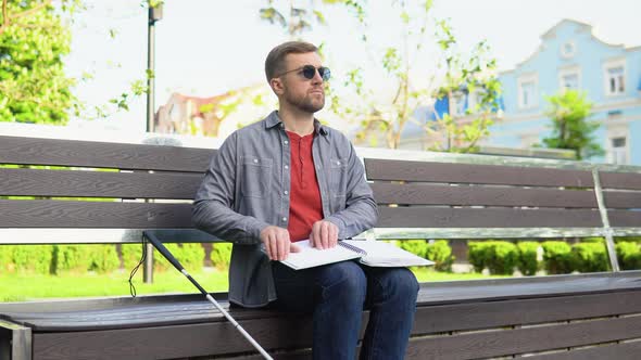 Blind Man Reading Braille Book Sitting on Bench in Summer Park