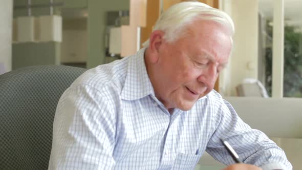 Senior Man Writing Memoirs In Book Sitting At Desk