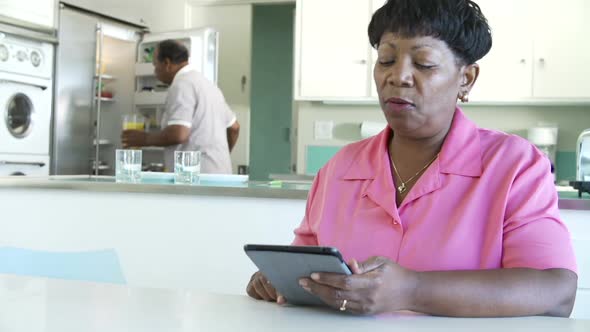 Senior Woman Using Digital Tablet As Husband Makes Breakfast 1