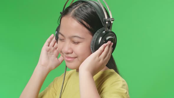 Side View Of Young Asian Kid Girl Listening To Music With Headphones And Dancing In The Green Screen alt