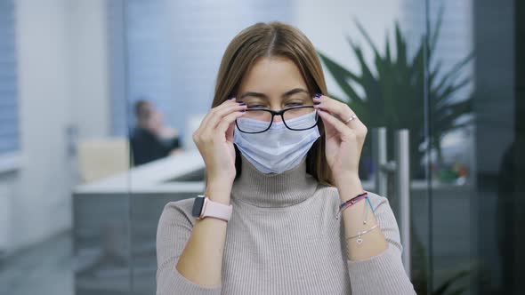 Woman in Protective Mask Puts on Glasses in the Office Looking Into the Camera Worker