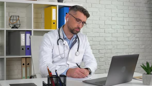 Doctor consults patient online video call laptop, writes symptoms in notebook