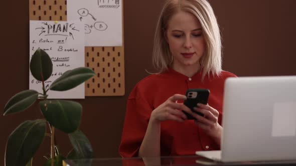 Blond hair woman in red shirt working at home office on laptop computer and speaking by mobile phone