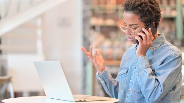 African Woman with Laptop Talking on Smartphone at Cafe 