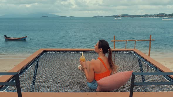 Beautiful Young Woman Sitting in Hammock Drinking Cocktail or Orange Juice on Sea Beach