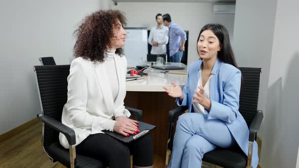 Positive Asian and Caucasian Women Talking and Shaking Hands in Office with Blurred Middle Eastern