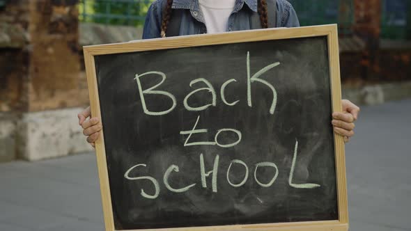 Hands of Little Girl Standing Holding and Raising Schoolboard with Text Back to School on the Street