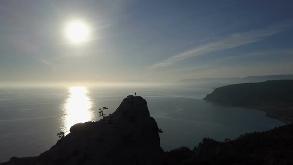 Aerial Silhouette of Young Woman on the Top of a Mountain Facing the Sea. Flight Over Lady on the