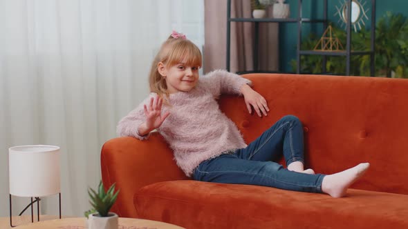 Child Girl Lying on Sofa at Home Looking at Camera Smiling Waving Hands Gesturing Hello or Goodbye