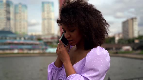 a Young African Woman Is Dressed in a Lilac Top with Ties and Puffy Sleeves. She Walks Along the