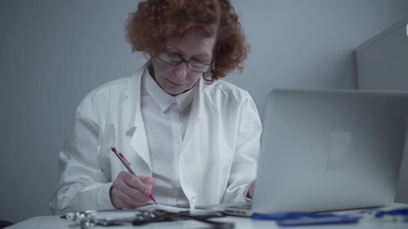 A Female Senior Doctor Makes a Medical Report Makes Notes in Medical Documents with a Pen While