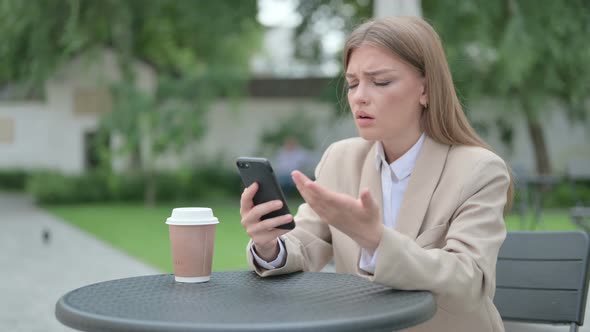 Young Businesswoman Reacting to Loss on Smartphone in Outdoor Cafe