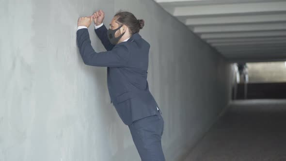 Side View of Angry Young Businessman in Covid-19 Face Mask Hitting Urban Underground Crossing Wall