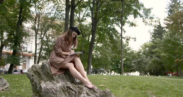 Girl Artist Sitting on a Stone in the Park with a Notebook and a Pencil in Her Hands