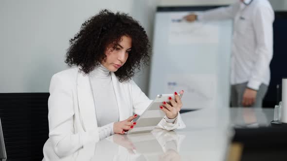 Concentrated Caucasian Curlyhaired Woman Using Tablet As Blurred Man Presenting Startup at