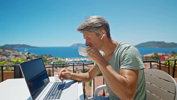 Dolly Zoom Out Footage Man in Earphones Working with Laptop Outdoors By Beautiful Blue Sea View