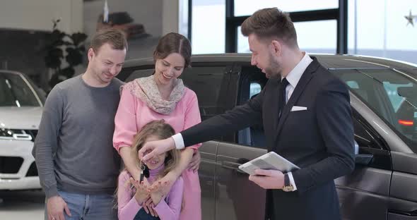 Cute Caucasian Little Girl Taking Car Keys From Dealer, Father Raising Her Up. Four People Looking