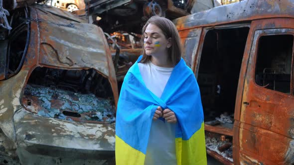 Young Sad Thoughtful Ukrainian Woman in National Flag Standing at Burnt Down Bombed Cars in War Zone