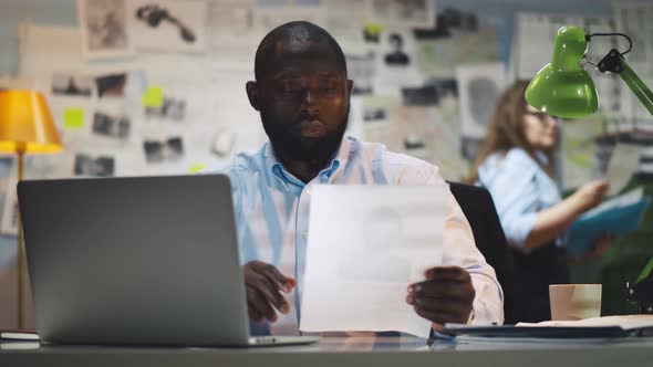 Portrait of Afro-american Policeman Studying Identikit of Criminal Sitting in Office