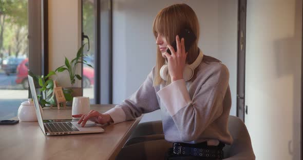 Young Busy Blond Student Female Talking on the Phone and Looking at Computer of the Modern Cozy Cafe