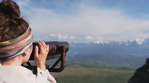 Attractive Girl Tourist on the Observation Deck Looks at the Mountains Through Binoculars