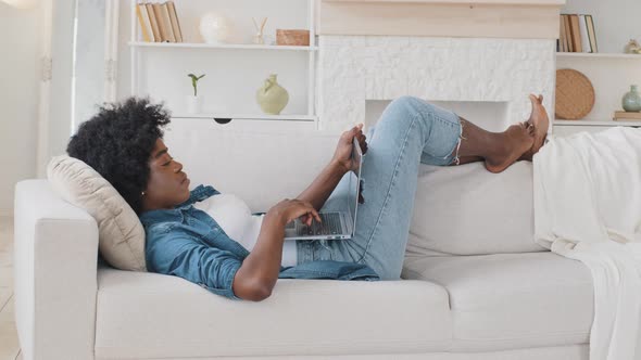 Smiling Serene African Young Woman Lying Down on Sofa Texting on Laptop