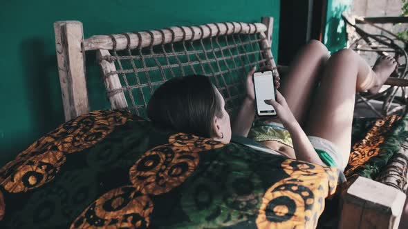 Young Girl Read Book From Smartphone Lying on Tropic Veranda of African Bungalow