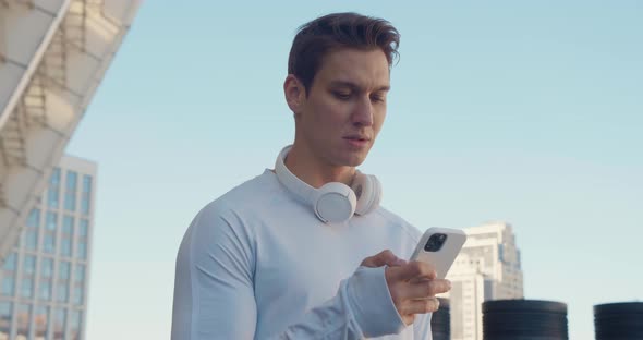 Athletic Young Man with Headphones Using Smartphone While Standing Outdoors at Stadium Before