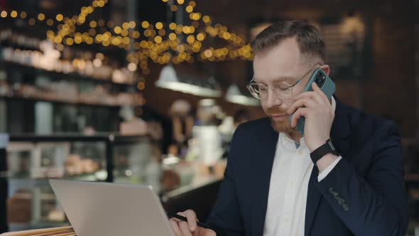 Businessman Talking on Mobile Phone Looking at Laptop While Working at Cafe